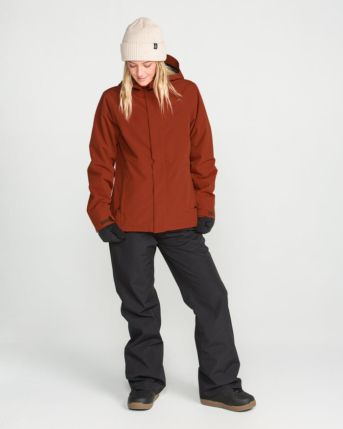 Wearing the Volcom Women’s Bolt Insulated Jacket in rust, a person pairs it with a beige beanie, black gloves, and black pants while standing against a plain white background, looking down with a relaxed posture.