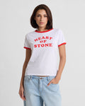 A woman with straight brown hair wears the Volcom Women's Brk Up Ringer Tee, a fitted top with red trim and bold HEART OF STONE lettering, paired with light blue jeans. She stands against a plain background with one hand in her pocket.