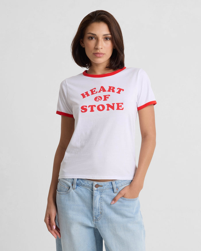 A woman with straight brown hair wears the Volcom Women's Brk Up Ringer Tee, a fitted top with red trim and bold HEART OF STONE lettering, paired with light blue jeans. She stands against a plain background with one hand in her pocket.