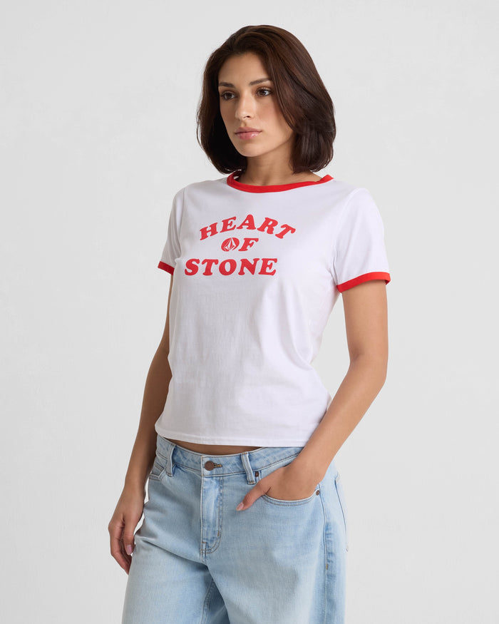 A woman with short brown hair wears the Volcom Women's Brk Up Ringer Tee—white with red trim and bold "HEART OF STONE" text—paired with light blue jeans, standing against a plain light background.