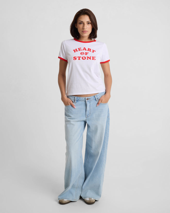 A woman faces forward in a Volcom Women's Brk Up Ringer Tee with red trim and "HEART OF STONE" text, paired with light blue wide-leg jeans and beige shoes, set against a plain light background.