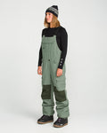 A person wearing a black long-sleeve shirt, Volcom Women’s Creston 3D Stretch Bib overalls in olive green with dark knee patches, dark snow boots, and a black beanie stands facing left against a plain white background.