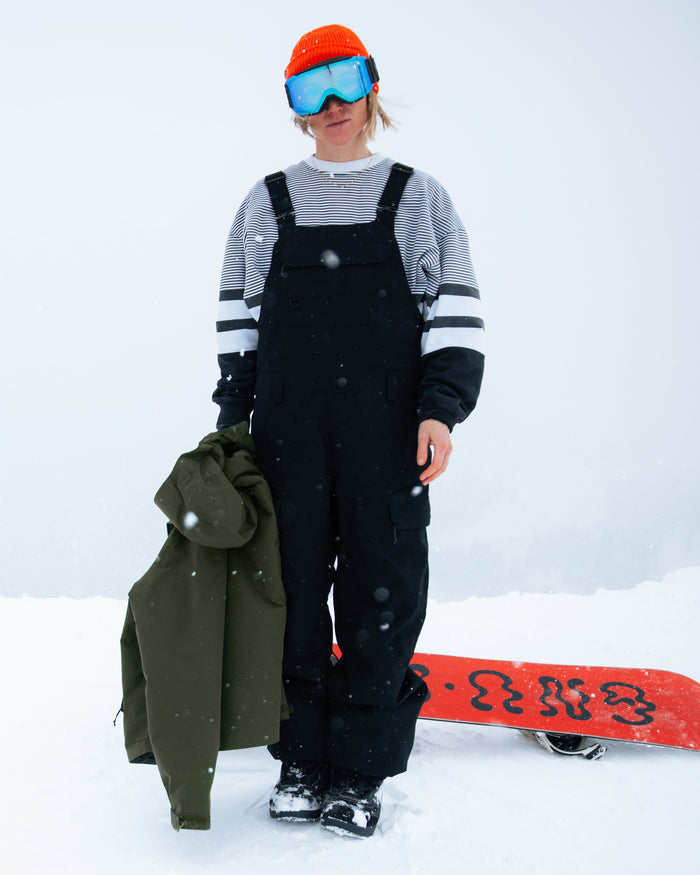 A person stands in the snow wearing Volcom’s Women’s Creston 3D Stretch Bib—black snow overalls with fully taped seams—and a striped shirt, holding a green jacket. They complete their look with a red helmet, blue goggles, and have a red snowboard on the ground beside them.