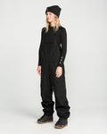Wearing Volcom’s Women’s Creston 3D Stretch Bib made from recycled stretch oxford fabric, a black long-sleeve shirt, black beanie, and gray boots, a person stands against a plain light background, facing slightly to the left and smiling.