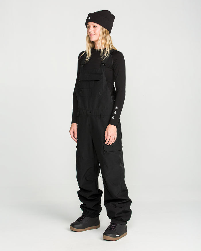 Wearing Volcom’s Women’s Creston 3D Stretch Bib made from recycled stretch oxford fabric, a black long-sleeve shirt, black beanie, and gray boots, a person stands against a plain light background, facing slightly to the left and smiling.