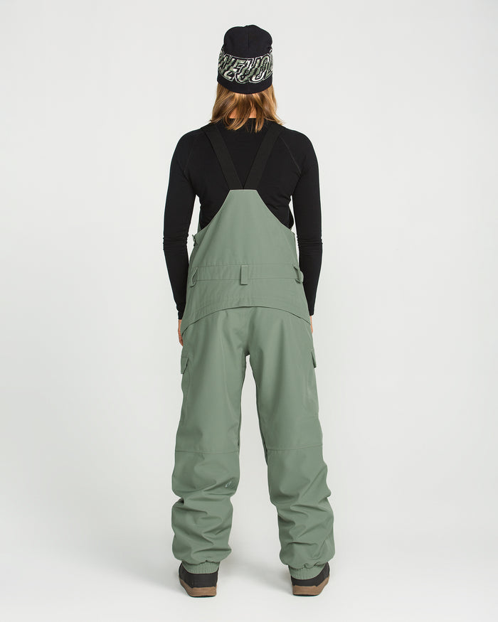 A person stands with their back to the camera, wearing Volcom Women’s Creston 3D Stretch Bib snow pants in sage green—crafted from recycled stretch oxford fabric with fully taped seams—paired with a black long-sleeve shirt, brown boots, and a black beanie featuring a white pattern, set against a plain white background.