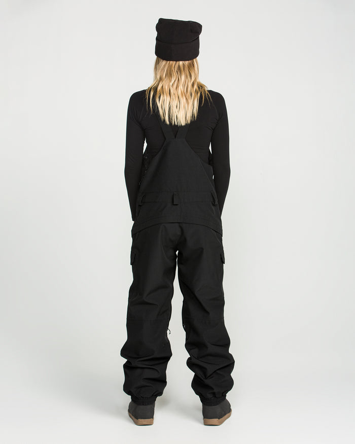 A person with long blonde hair, wearing a black beanie, a black long-sleeve shirt, and Volcom Women’s Creston 3D Stretch Bib overalls—crafted from recycled stretch oxford fabric—stands facing away against a plain white background.