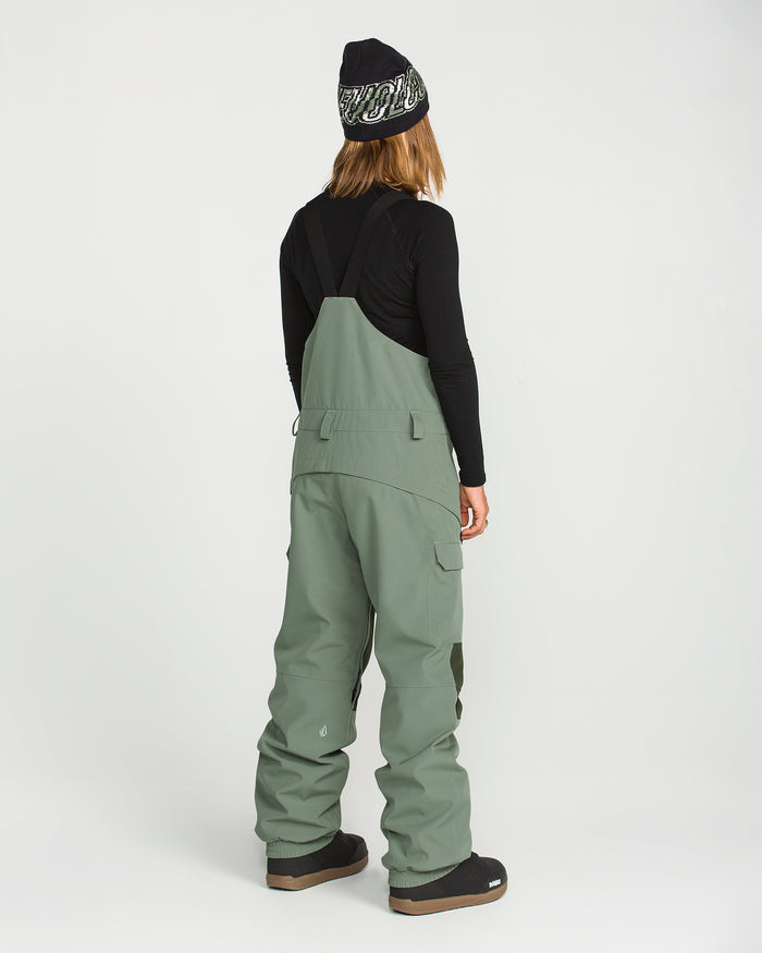 A person stands with their back to the camera, dressed in a black long-sleeve shirt and the Volcom Women’s Creston 3D Stretch Bib in sage green, made from recycled stretch oxford. The look is completed with black boots and a black beanie featuring white lettering, set against a plain white background.
