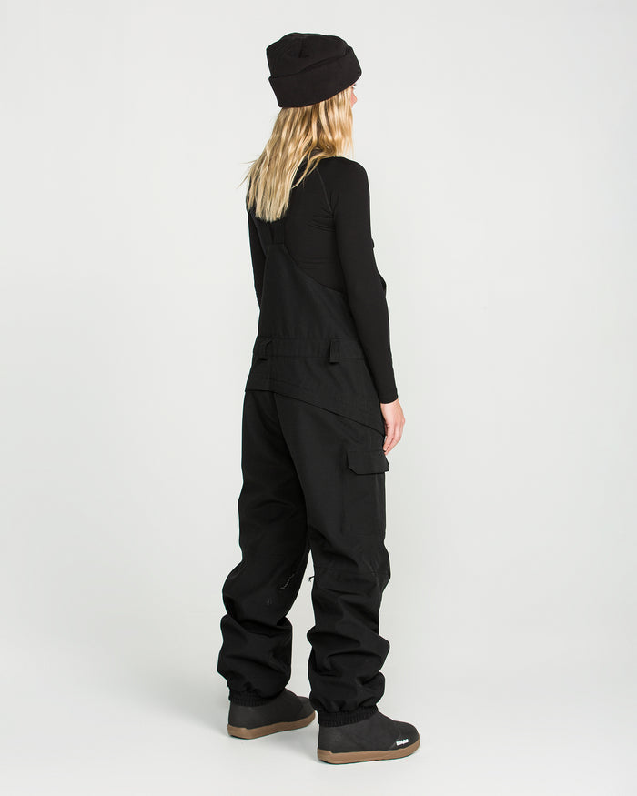 A person with long blonde hair, wearing a black beanie, black long-sleeve top, and the Volcom Women’s Creston 3D Stretch Bib—crafted from recycled stretch oxford fabric with fully taped seams—stands facing away on a plain white background.