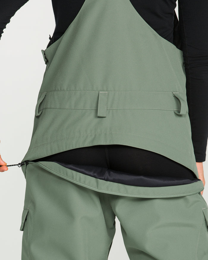 A person is shown from the lower torso with hands visible, unzipping the back of Volcom’s Women’s Creston 3D Stretch Bib—green overalls crafted from recycled stretch oxford fabric—to reveal black clothing underneath.