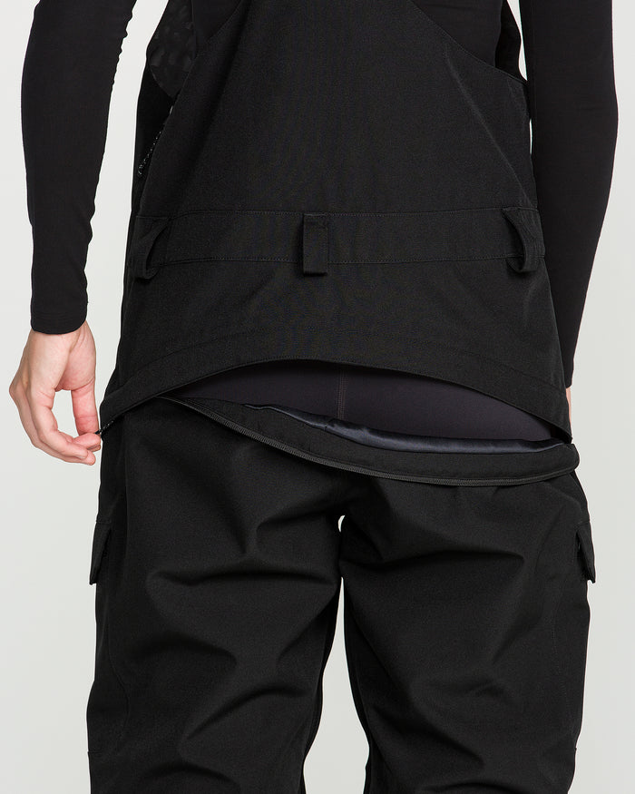 A person is shown from behind wearing the Volcom Women’s Creston 3D Stretch Bib, crafted from recycled stretch oxford fabric, paired with a jacket. The back flap of the jacket is partially lifted, revealing the inner waistband area of the bib pants. The background is plain and light-colored.