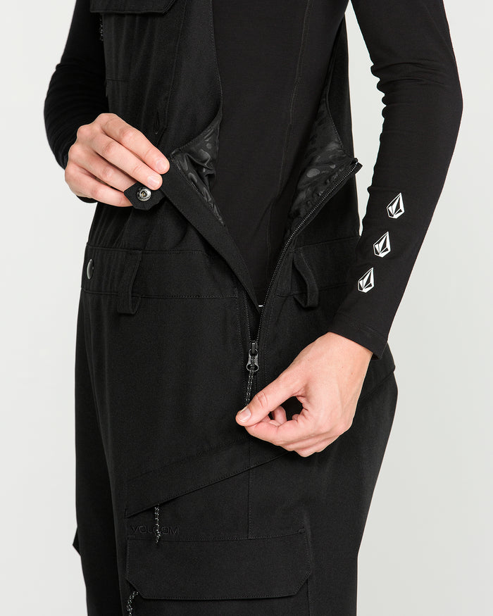 A person, visible only by their torso and hands, zips up a black Volcom Women’s Creston 3D Stretch Bib made from recycled stretch oxford. The black long-sleeve garment features three white embroidered geometric logos on the sleeve.