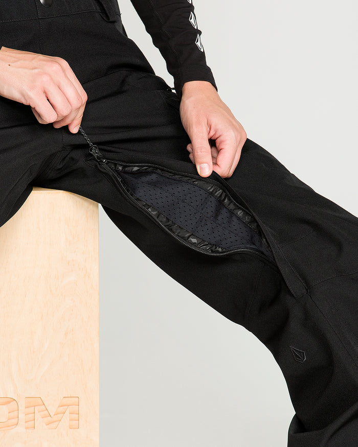 A person sitting on a wooden block unzips a vent on the inner thigh of the Volcom Women’s Creston 3D Stretch Bib, revealing a mesh lining inside.