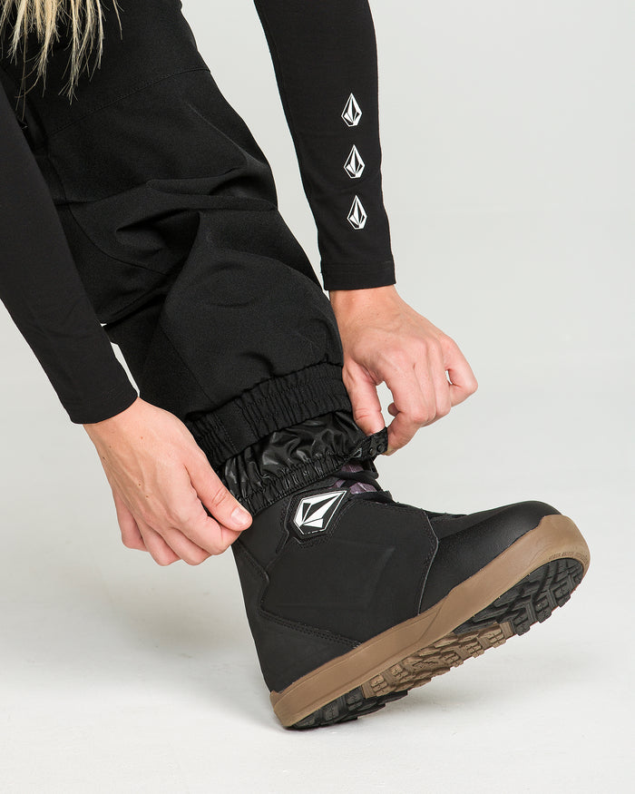 A person adjusts the Volcom Women’s Creston 3D Stretch Bib, made from recycled stretch oxford fabric in black, over a black snow boot with a brown sole. Both the bib pants and the boot feature a white geometric Volcom logo. Hands are visible against a plain, light-colored background.