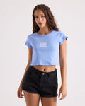 A woman with long curly hair wears the Volcom Women's Detour 1991 Mid Tee—a blue, 100% cotton, cropped fitted t-shirt with white text—paired with black denim shorts, standing against a plain light background.