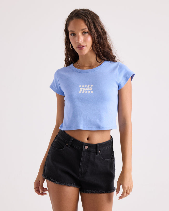 A woman with long curly hair wears the Volcom Women's Detour 1991 Mid Tee—a blue, 100% cotton, cropped fitted t-shirt with white text—paired with black denim shorts, standing against a plain light background.