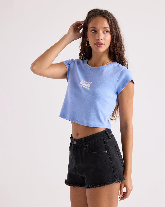 A woman with long curly hair wears the Volcom Women's Detour 1991 Mid Tee in light blue, a 100% cotton cropped fitted style, paired with black denim shorts. She stands against a plain light background with one arm raised to her head.