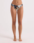 A woman models Volcom's Women's Distant Tropic Hipster Cheekini bikini bottoms, featuring a tropical print and high-cut leg, standing barefoot against a white background with arms relaxed at her sides, visible from shoulders to feet.