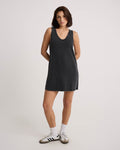 A woman with medium-length brown hair wears the Volcom Women's Glam Crop Dress, a sleeveless mini in Vintage Black, paired with white Adidas sneakers featuring black stripes, standing hands behind her back against a plain light backdrop.