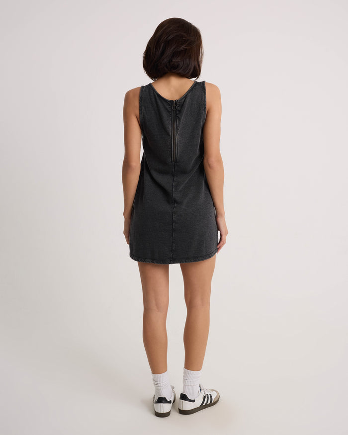 A woman with shoulder-length dark hair faces away, wearing the Volcom Women's Glam Crop Dress in Vintage Black with a visible back zipper, paired with white socks and black-and-white sneakers against a plain background.