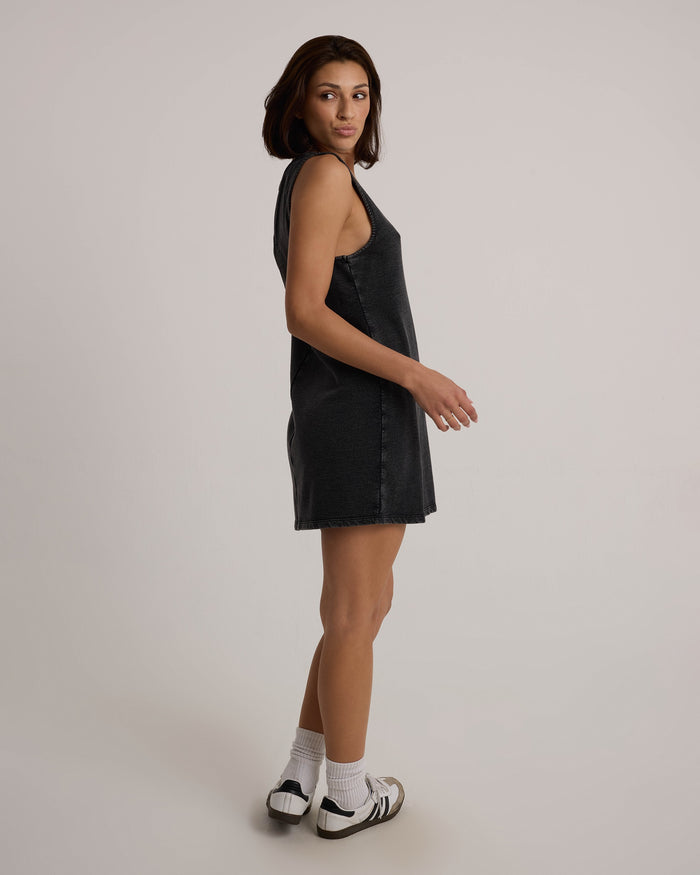 A woman with shoulder-length brown hair models the Volcom Women's Glam Crop Dress, a sleeveless dark denim mini, styled with white socks and sneakers. She stands sideways and glances back, evoking a subtle vintage black vibe against a plain background.