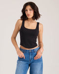 A woman with short dark hair models the Volcom Women's Hot Girl fitted black tank top and blue jeans, posing with one hand in her pocket against a plain light background.