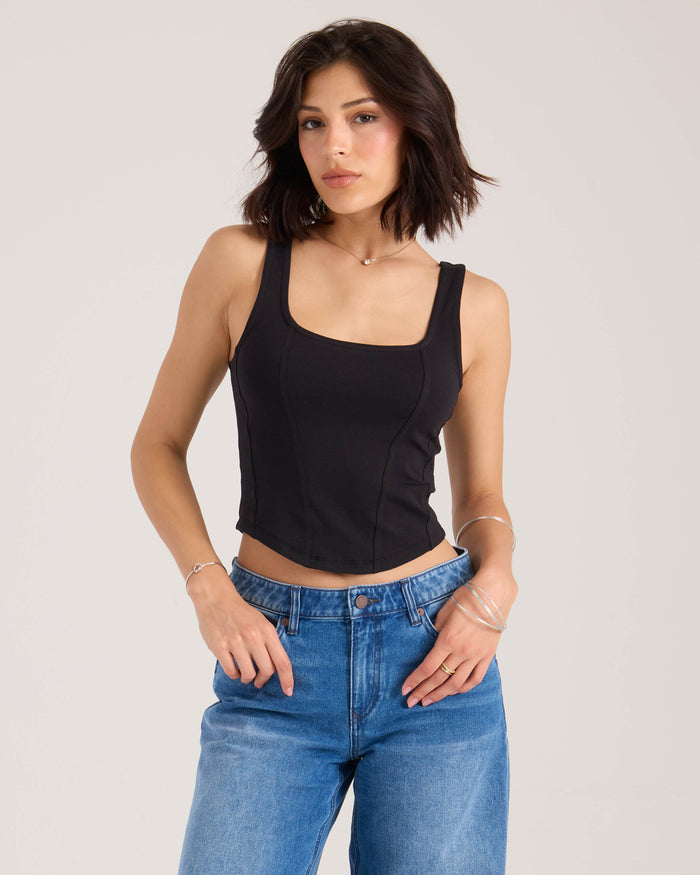 A woman with short dark hair models the Volcom Women's Hot Girl fitted black tank top and blue jeans, posing with one hand in her pocket against a plain light background.