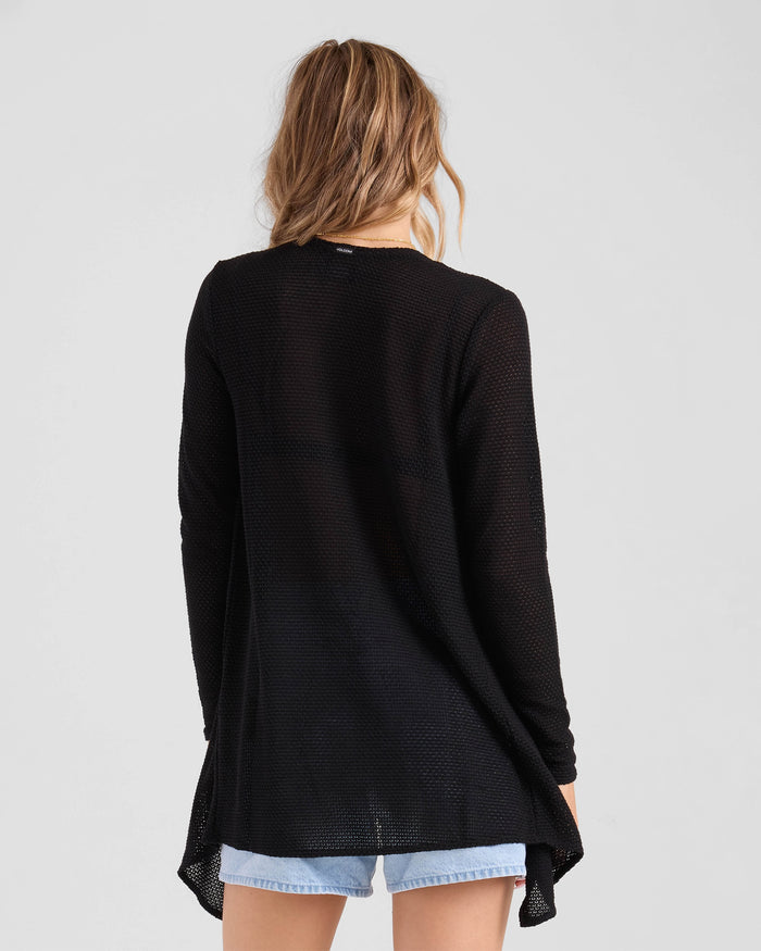 A woman with wavy, light brown hair is seen from the back wearing Volcom's Women's Lived In Go Go Wrap cardigan with light blue denim shorts against a plain light background.