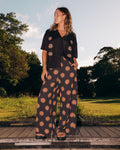 A person with wavy hair stands outdoors wearing a loose black shirt and Volcom Women's Midnite Ramble Pants, which are high-waisted with a tan starburst pattern, paired with black sandals amid green trees under a partly cloudy sky.