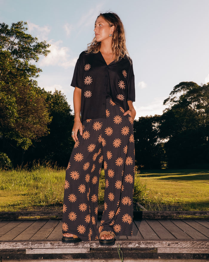 A person with wavy hair stands outdoors wearing a loose black shirt and Volcom Women's Midnite Ramble Pants, which are high-waisted with a tan starburst pattern, paired with black sandals amid green trees under a partly cloudy sky.