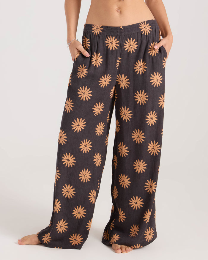 A person stands barefoot wearing Volcom Women's Midnite Ramble Pant—high-waisted, black wide-leg pants with an orange daisy print. Their hands are in the pockets, and only their torso and legs are visible.