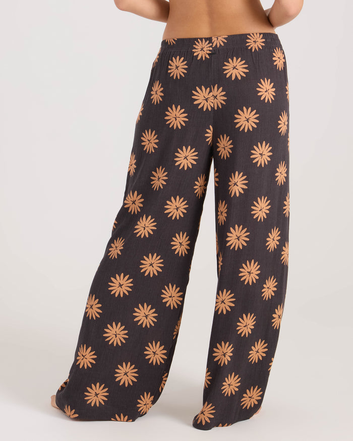 A woman wears Volcom's Women's Midnite Ramble Pant, a high-waisted, wide-leg black pant with an all-over orange daisy floral design, standing barefoot against a plain light background.