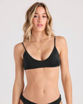 A woman with wavy blonde hair wears the Volcom Women's Simply Seamless Bralette, a black bikini top made from recycled nylon, along with a gold necklace. She stands against a plain light background facing forward with a neutral expression.