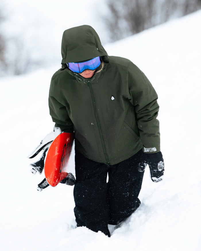 Wearing the Volcom Women’s Steele Insulated Jacket, a person in green with goggles walks uphill in the snow, holding a red sled and carrying a snowboard under one arm.