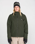 A person is wearing the Volcom Women’s Steele Insulated Jacket in dark green, paired with beige pants, black gloves, and a patterned black beanie, standing facing forward against a plain light background.