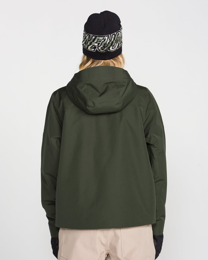 A person is facing away from the camera against a plain white background, wearing the Volcom Women’s Steele Insulated Jacket in dark green, paired with black gloves, beige pants, and a black beanie with white text. The jacket features recycled insulation.