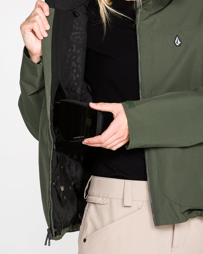 A woman wearing the Volcom Women’s Steele Insulated Jacket in green, featuring recycled insulation, and beige pants displays the inside pocket while holding a pair of ski goggles.