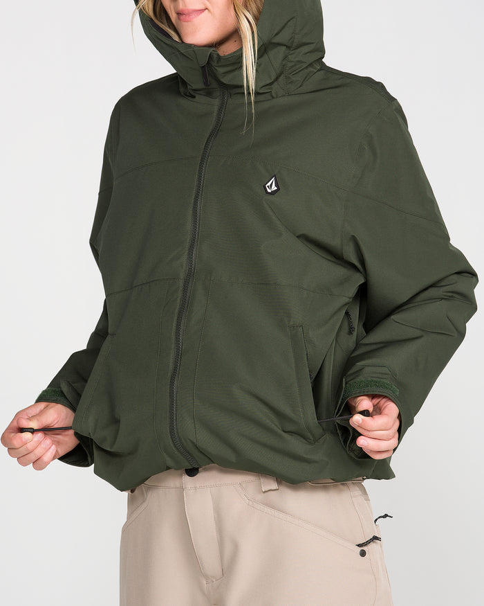 A person wearing the Volcom Women’s Steele Insulated Jacket in dark green adjusts the drawstrings at the hem. The waterproof ski jacket, made with recycled insulation, features a chest logo and zippered pockets. The person, dressed in beige pants, has their face partially visible and turned to the side.