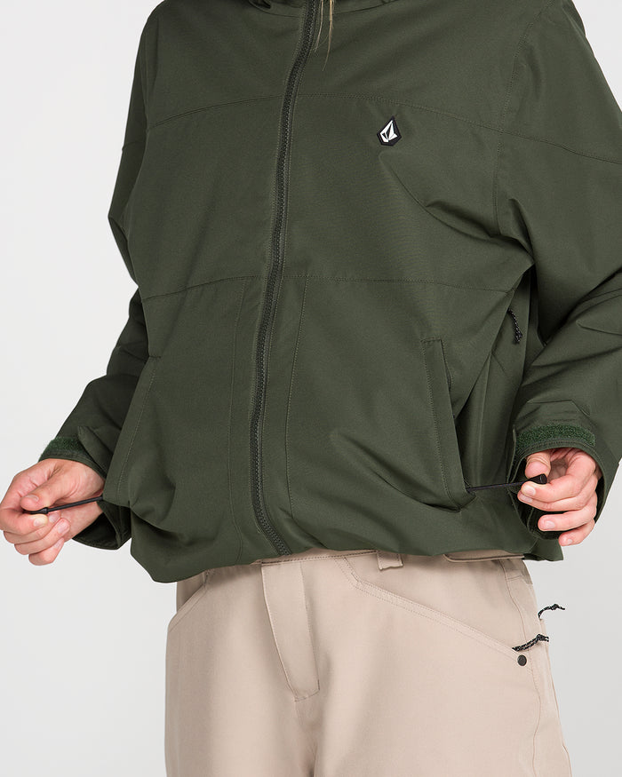 A person wearing the Volcom Women’s Steele Insulated Jacket in green and beige pants adjusts a drawstring at the hem of the jacket, pulling it tight with both hands. The waterproof ski jacket features zippered pockets, recycled insulation, and a small Volcom logo on the chest. The person’s face is not shown.