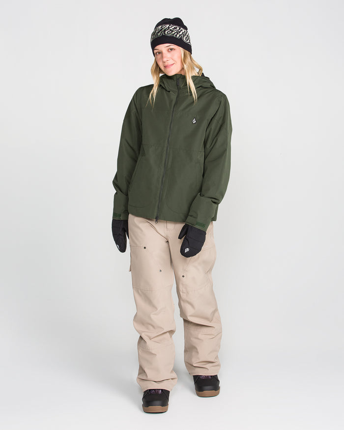 A person wearing a black beanie, black gloves, beige cargo pants, black shoes, and the Volcom Women’s Steele Insulated Jacket—a green waterproof ski jacket with recycled insulation—stands against a plain white background, smiling.