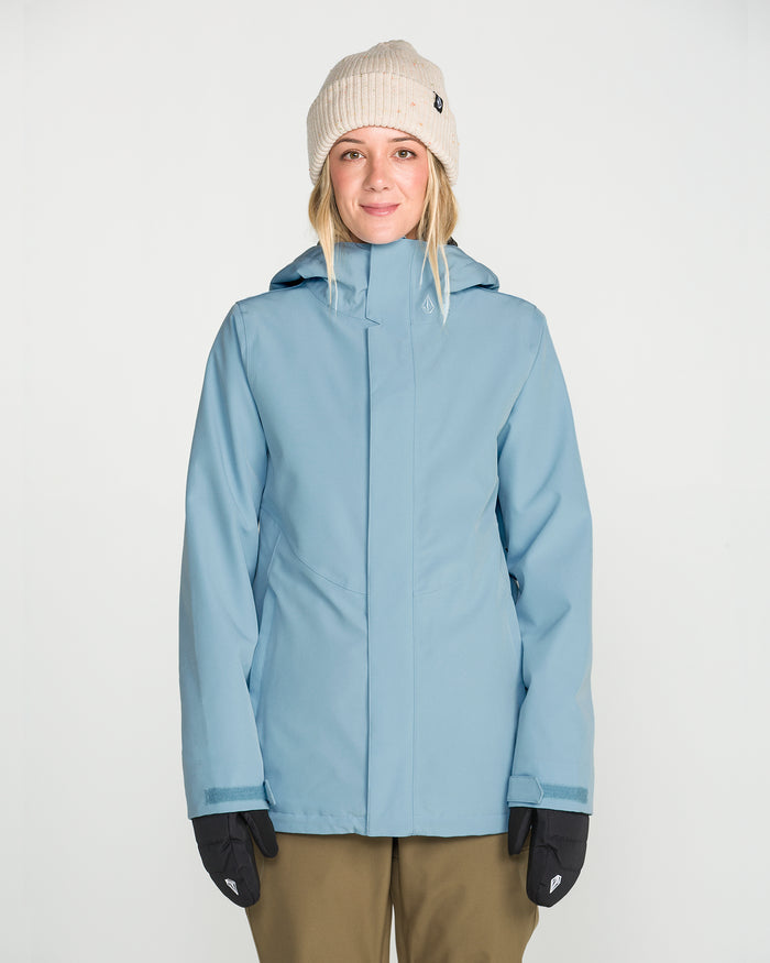 A person wearing the Volcom Women’s Stoney Shadow Insulated Jacket in light blue, styled with a beige knit beanie, black gloves, and olive green pants, stands facing the camera against a plain white background.