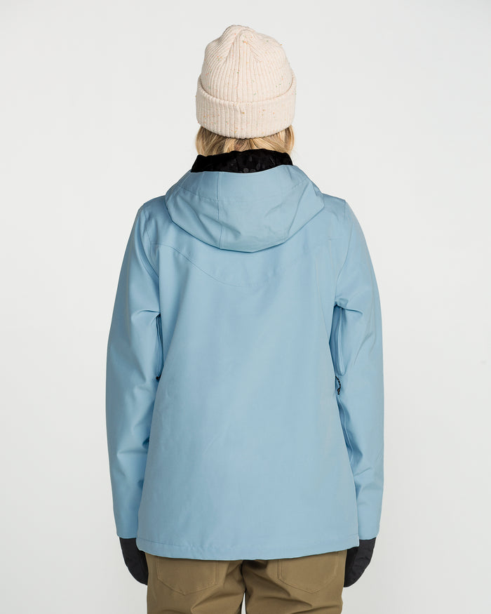 A person facing away, wearing the Volcom Women’s Stoney Shadow Insulated Jacket in light blue with recycled insulation, paired with a beige knit beanie, black gloves, and tan pants against a plain white background.