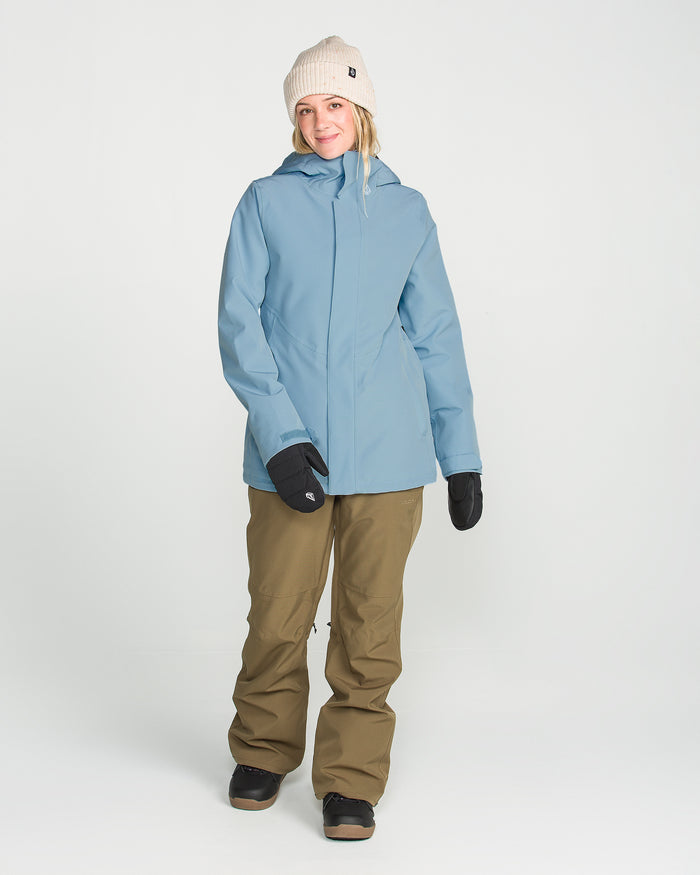 A woman wearing the Volcom Women’s Stoney Shadow Insulated Jacket in light blue—featuring waterproof construction and recycled insulation—pairs it with a beige beanie, black gloves, brown pants, and black shoes as she stands against a plain white background, smiling slightly.