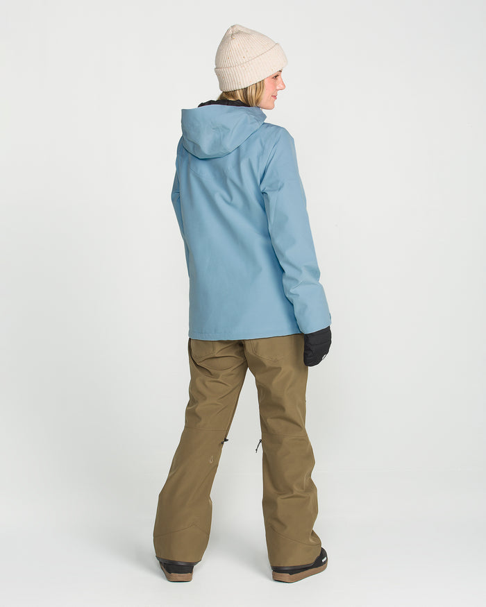A person wearing the Volcom Women’s Stoney Shadow Insulated Jacket in light blue, paired with a beige knit hat, brown snow pants, and black gloves, stands facing away from the camera against a white background.
