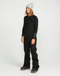 A person stands facing slightly left against a plain white background, wearing a gray beanie, black long-sleeve shirt, and the Volcom Women’s Swift Bib—sleek black women’s overalls made from V-Science stretch recycled fabric.