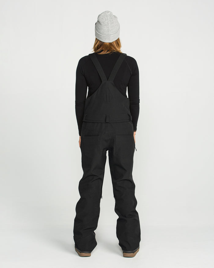A person stands facing away from the camera against a plain white background, wearing a gray beanie, black long-sleeve shirt, and the Volcom Women’s Swift Bib made from recycled fabric.
