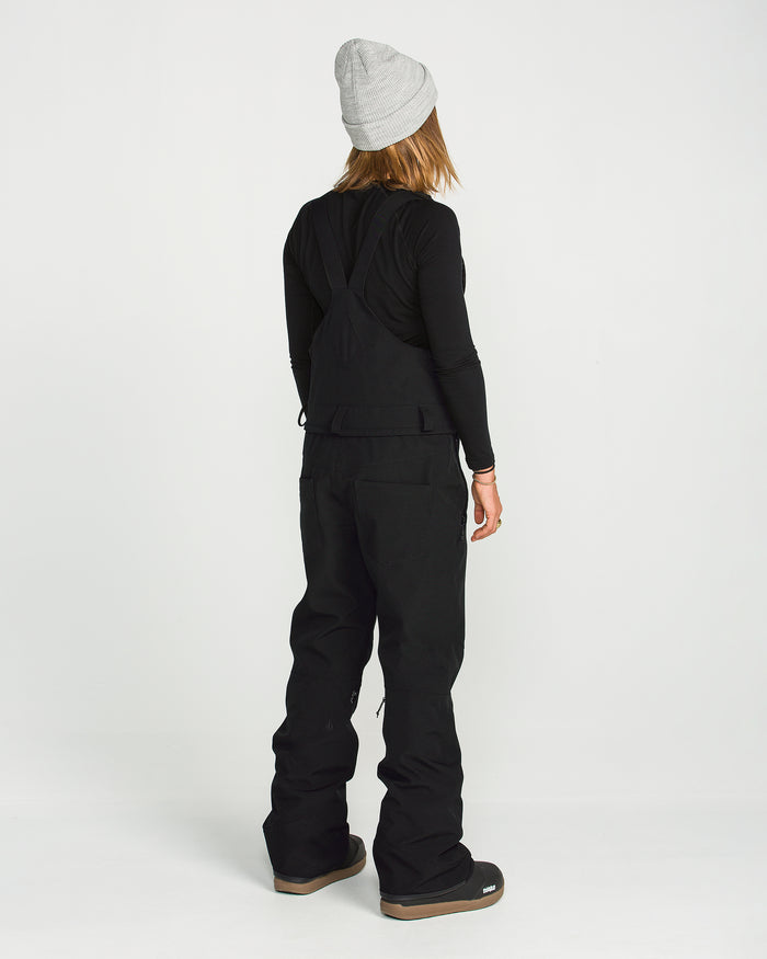 A person stands facing away, wearing a gray beanie, a black long-sleeve shirt, and the Volcom Women’s Swift Bib made from V-Science stretch recycled fabric, paired with black pants and brown-soled shoes against a plain white background.