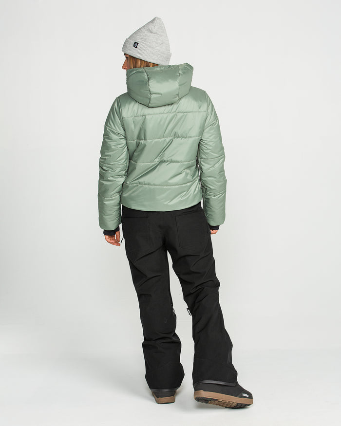 A person stands facing away, wearing the Volcom Women’s Swift Bib in black, paired with a light green puffer jacket made from recycled fabric, brown boots, and a gray beanie, against a plain white background.