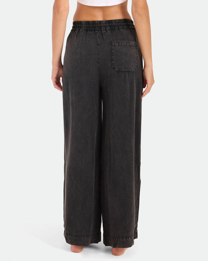 A person stands barefoot facing away, wearing Volcom Women's Washed Up Twill Pants—black wide-leg style with an elastic waist and a single back pocket—against a plain white background.