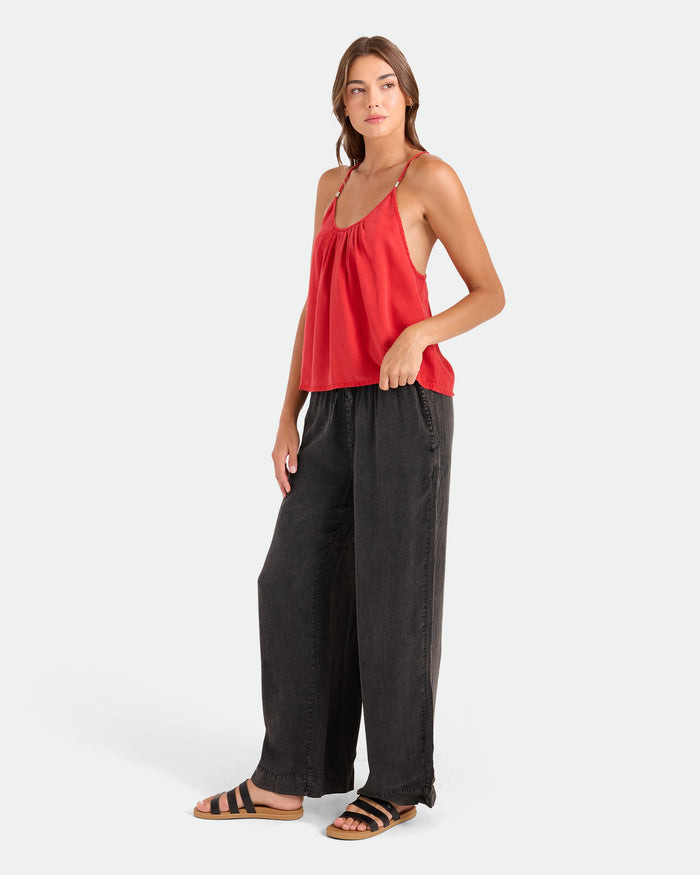 A woman with long brown hair poses against a plain background wearing Volcom Women's Washed Up Twill Pants, featuring a wide-leg silhouette and elastic waist, paired with a red camisole and black sandals, one hand in her pocket, looking to the side.